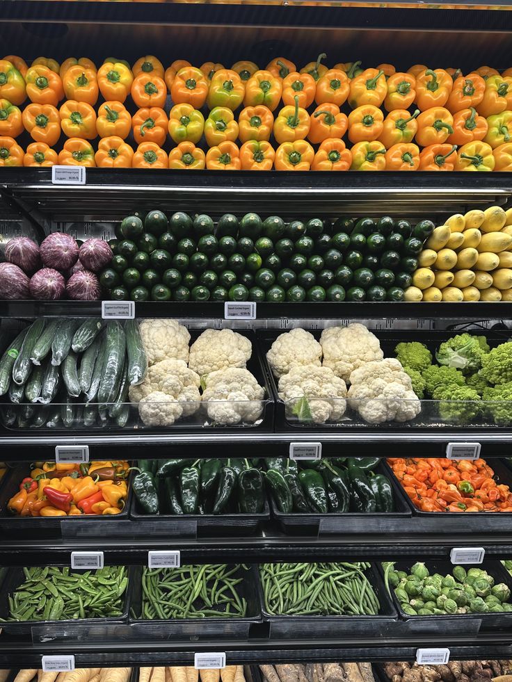 Assorted fresh vegetables on supermarket shelves, including peppers, cucumbers, cauliflower, and green beans.