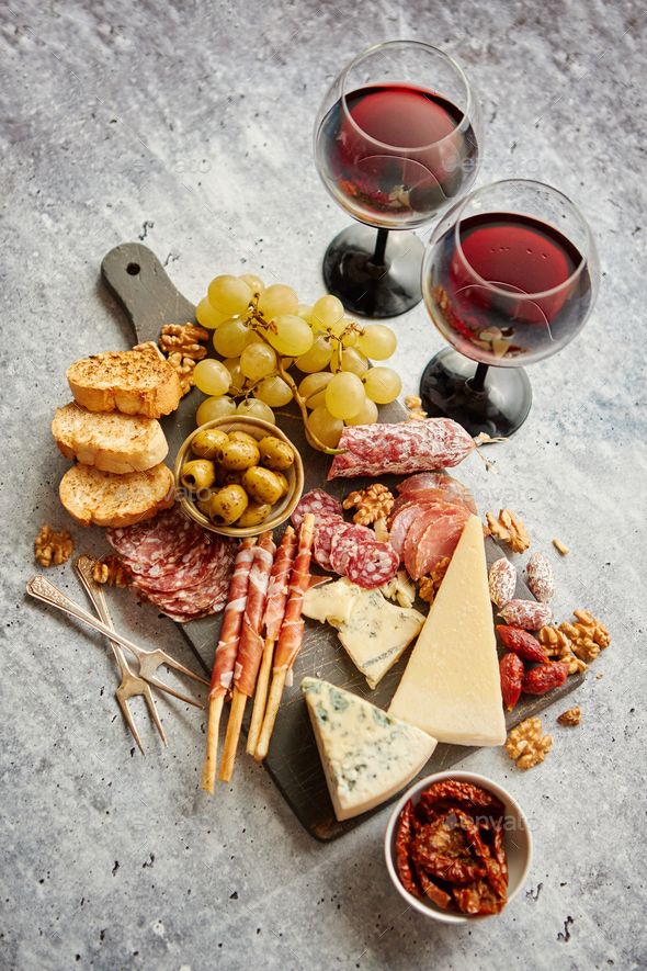 Cheese and charcuterie board with olives, grapes, cured meats, and two glasses of red wine on a textured surface.