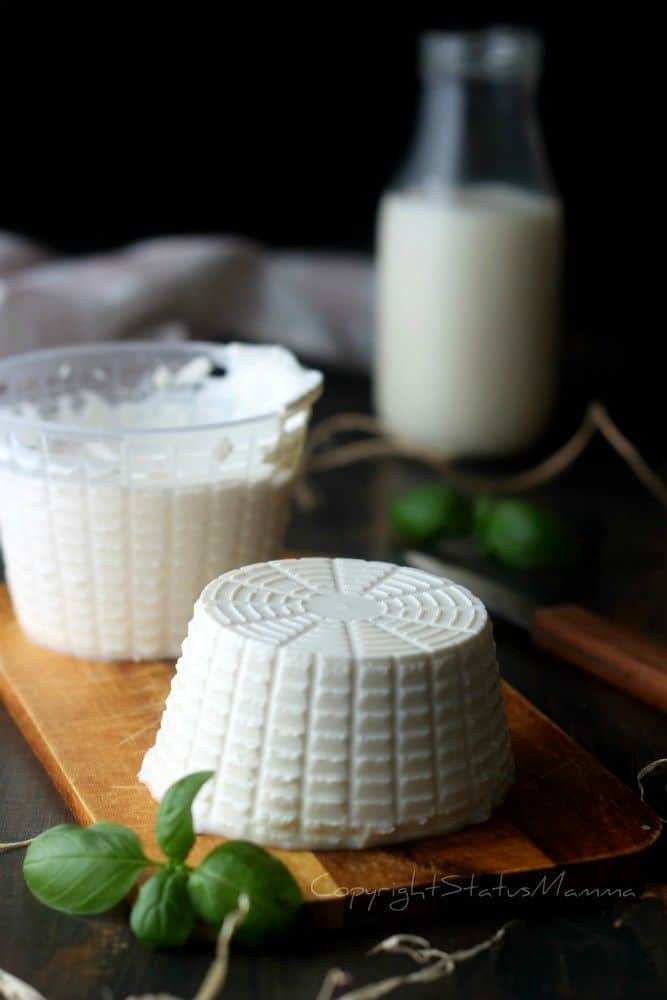 Fresh ricotta cheese on a wooden board with basil leaves and a bottle of milk in the background.