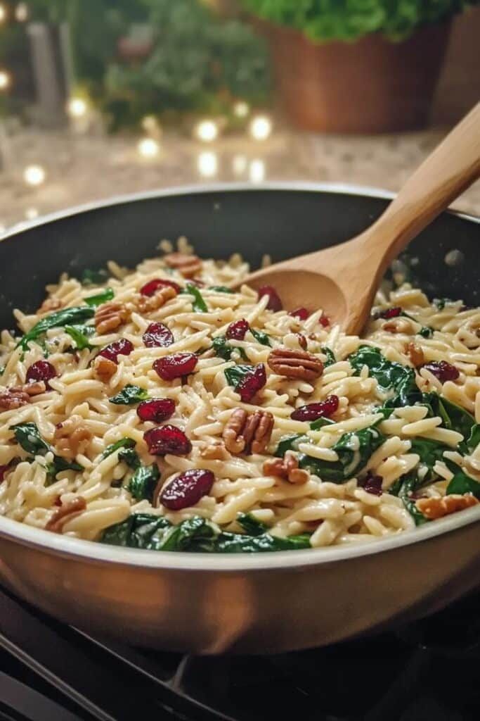 Creamy orzo pasta with spinach, pecans, and cranberries in a skillet, stirred with a wooden spoon.