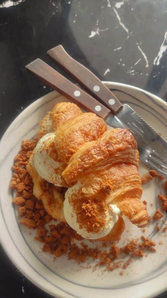 Croissant with ice cream and crumbled cookies on a plate, accompanied by a rustic knife and fork set.