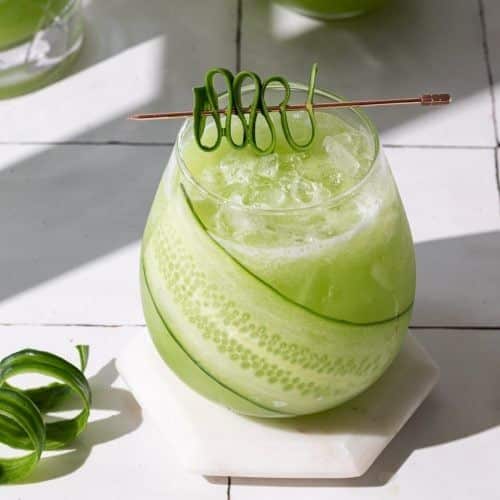 Green cucumber cocktail with ice and artistic garnish on a white tile background. Refreshing summer drink display.