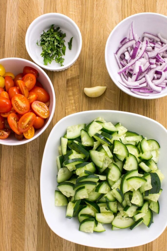 Chopped ingredients for a fresh salad: cucumbers, tomatoes, red onion, basil, and garlic on a wooden surface.