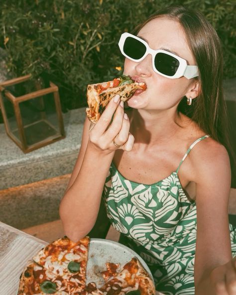 Woman in sunglasses enjoying a slice of pizza outdoors, wearing a green floral dress.