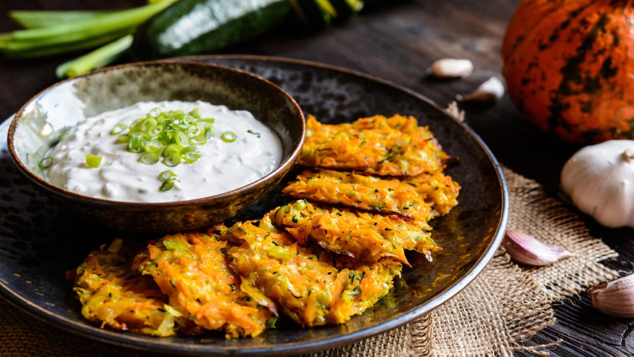 Crispy vegetable fritters served with creamy dip garnished with green onions on rustic wooden table.