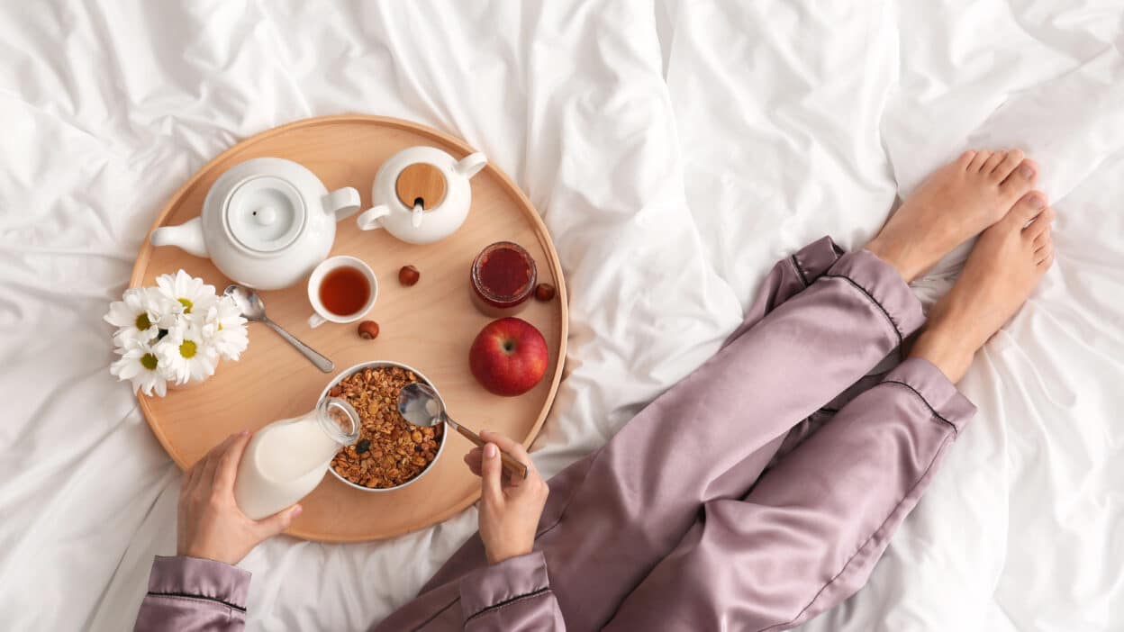 Breakfast in bed with granola, milk, tea, and an apple on a tray with flowers, next to person in lavender pajamas.