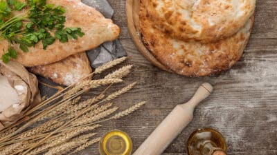 Freshly baked bread with herbs, flour, wheat, rolling pin, and olive oil on rustic wood table.