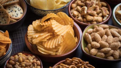 Variety of salty snacks in bowls, including potato chips, pretzels, peanuts, and crackers, on a dark table surface.