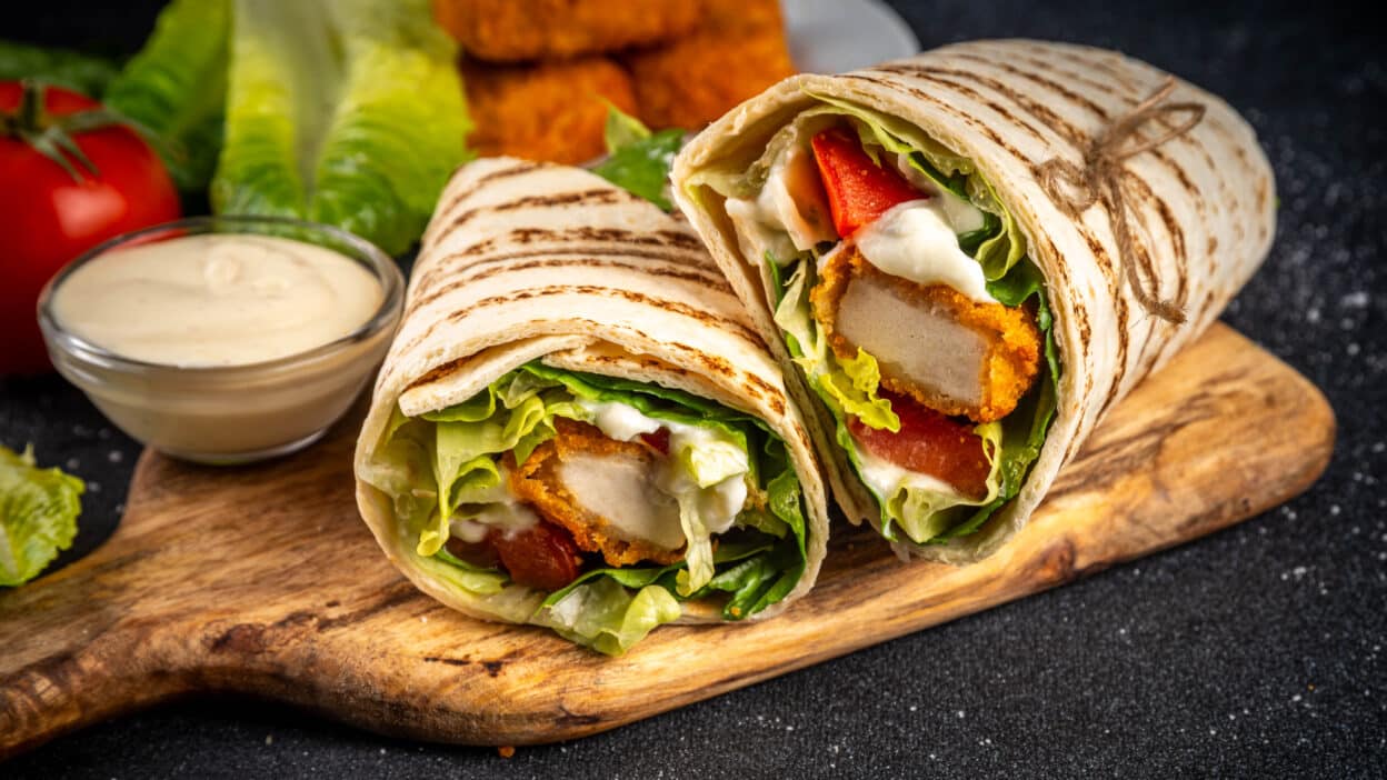 Delicious chicken wrap with lettuce, tomato, and creamy sauce on a wooden board, garnished with crispy chicken strips.