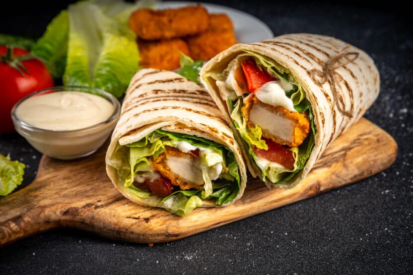 Delicious chicken wrap with lettuce, tomato, and creamy sauce on a wooden board, garnished with crispy chicken strips.