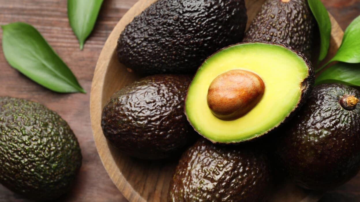 Ripe avocados in a wooden bowl, one cut in half, showcasing creamy texture and large seed, with green leaves around.