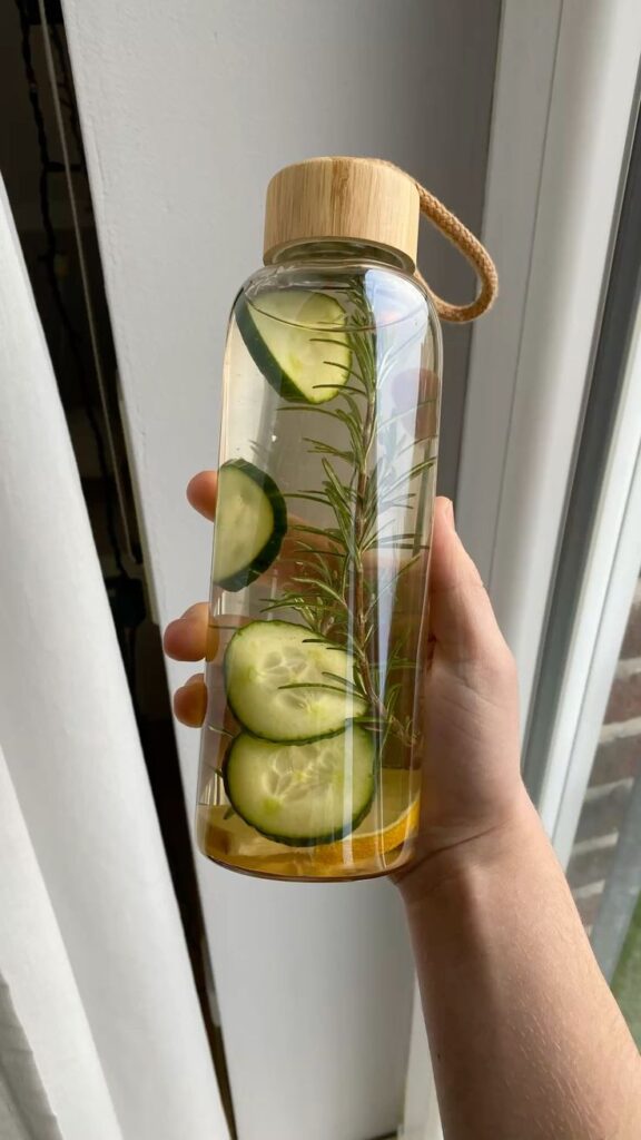 Infused water bottle with cucumber slices and rosemary, held by hand near a window.