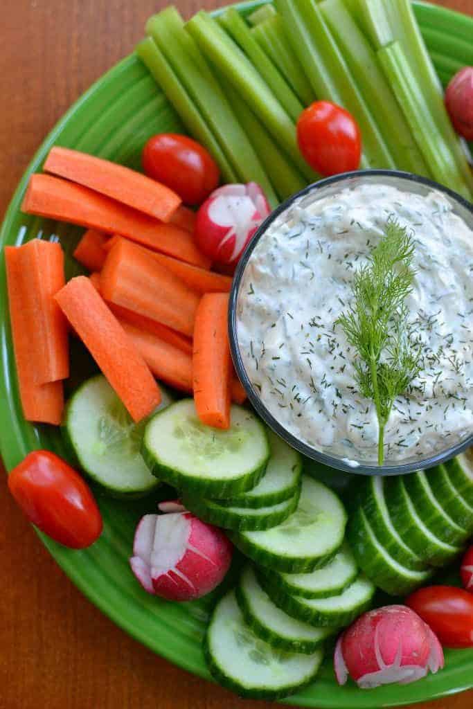 Veggie platter with carrots, cucumbers, celery, cherry tomatoes, radishes, and creamy dill dip on a green plate.