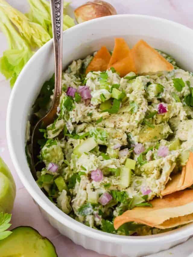 Chicken salad with avocado, red onion, and herbs in a white bowl, garnished with pita chips and a spoon.