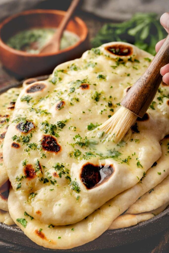 Brushing garlic butter on freshly baked naan bread, garnished with herbs, resting on a plate with sauce bowl nearby.