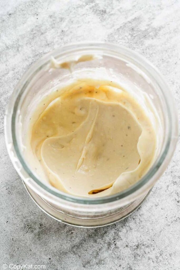 Creamy garlic aioli in a glass jar on a textured gray surface, perfect for sandwiches and dipping.