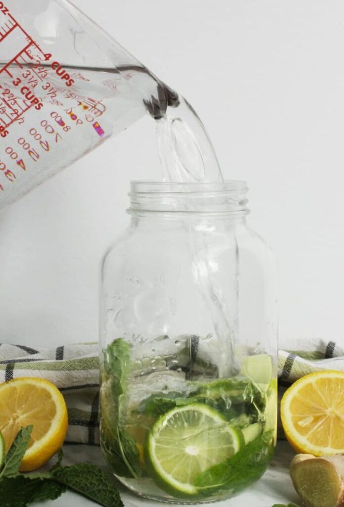 Pouring water into a mason jar with lime, lemon, mint, and ginger for a refreshing detox drink.