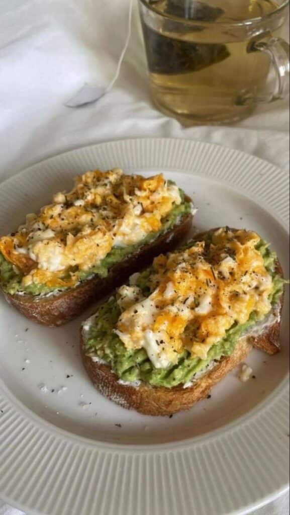 Avocado toast topped with scrambled eggs on a white plate, served with a cup of herbal tea.