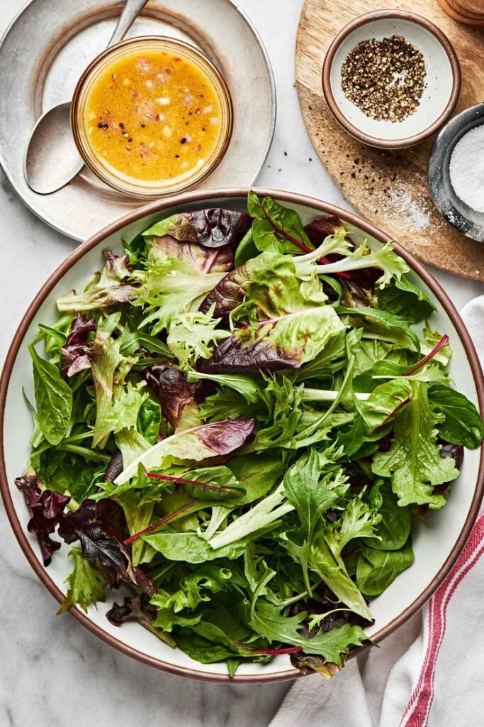 Fresh mixed green salad with mustard vinaigrette, salt, and pepper on a marble surface.