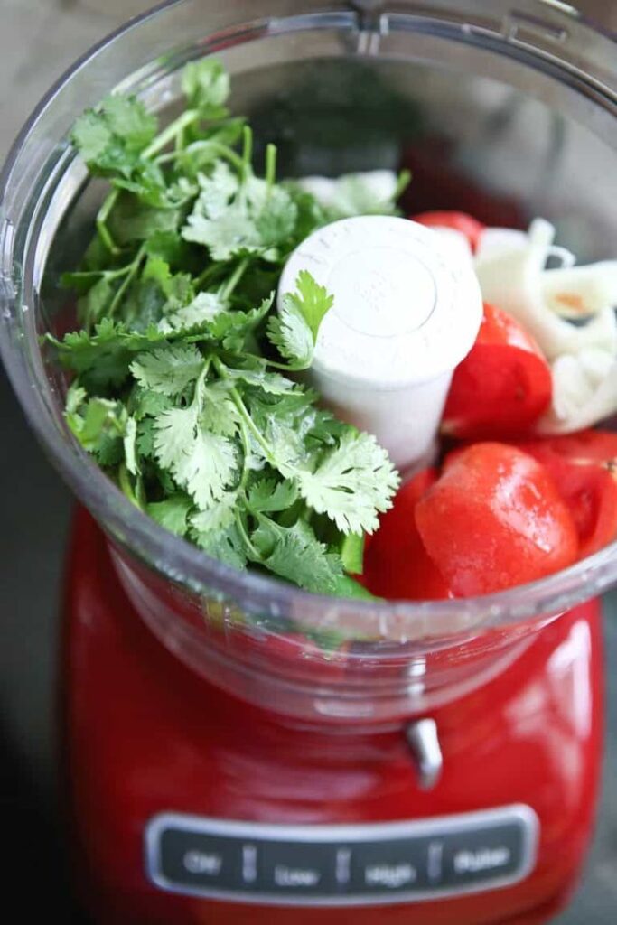 Fresh cilantro, tomatoes, and onions in a food processor ready for blending on a red base.