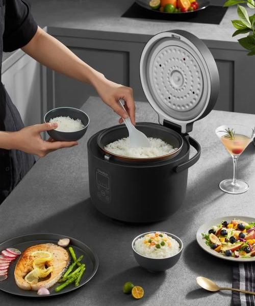 Person serving rice from a cooker, with plates of food and a drink on a kitchen countertop.