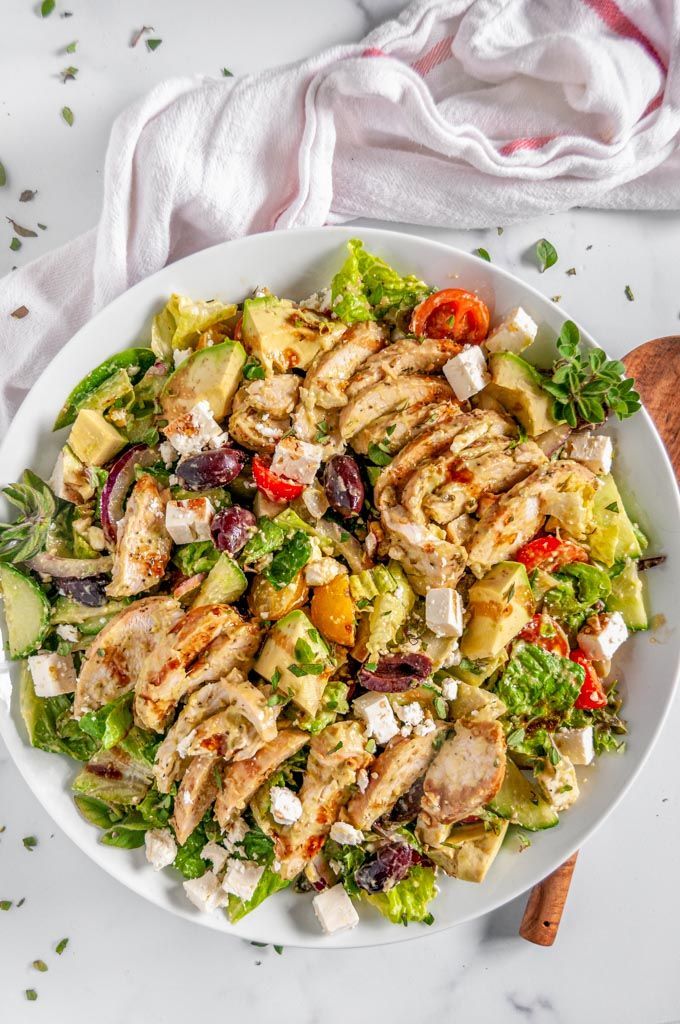 Fresh Greek salad with grilled chicken, avocado, feta, olives, tomato, and greens on a white plate.