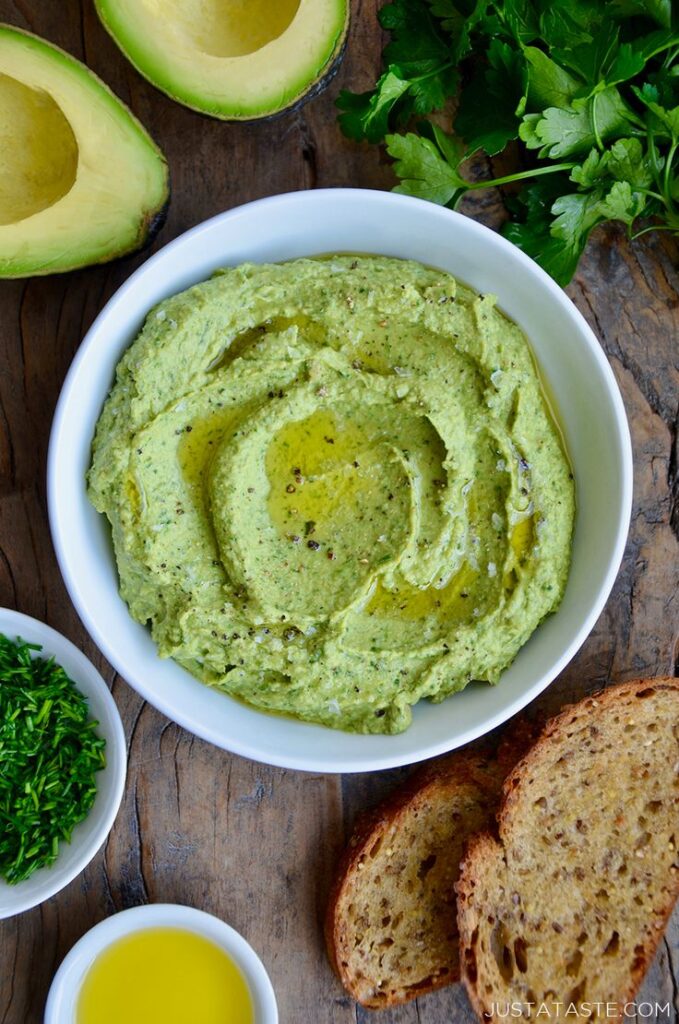 Creamy avocado hummus in a white bowl, garnished with olive oil and herbs, ready to serve with sliced bread.