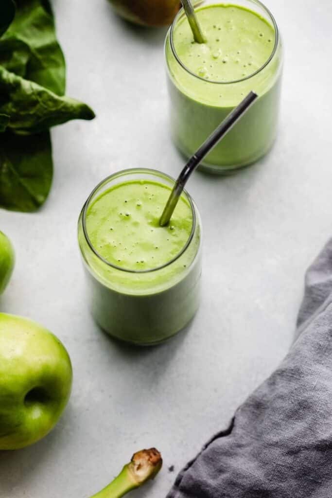 Green smoothies in glass jars with leafy greens and apples on a light background.