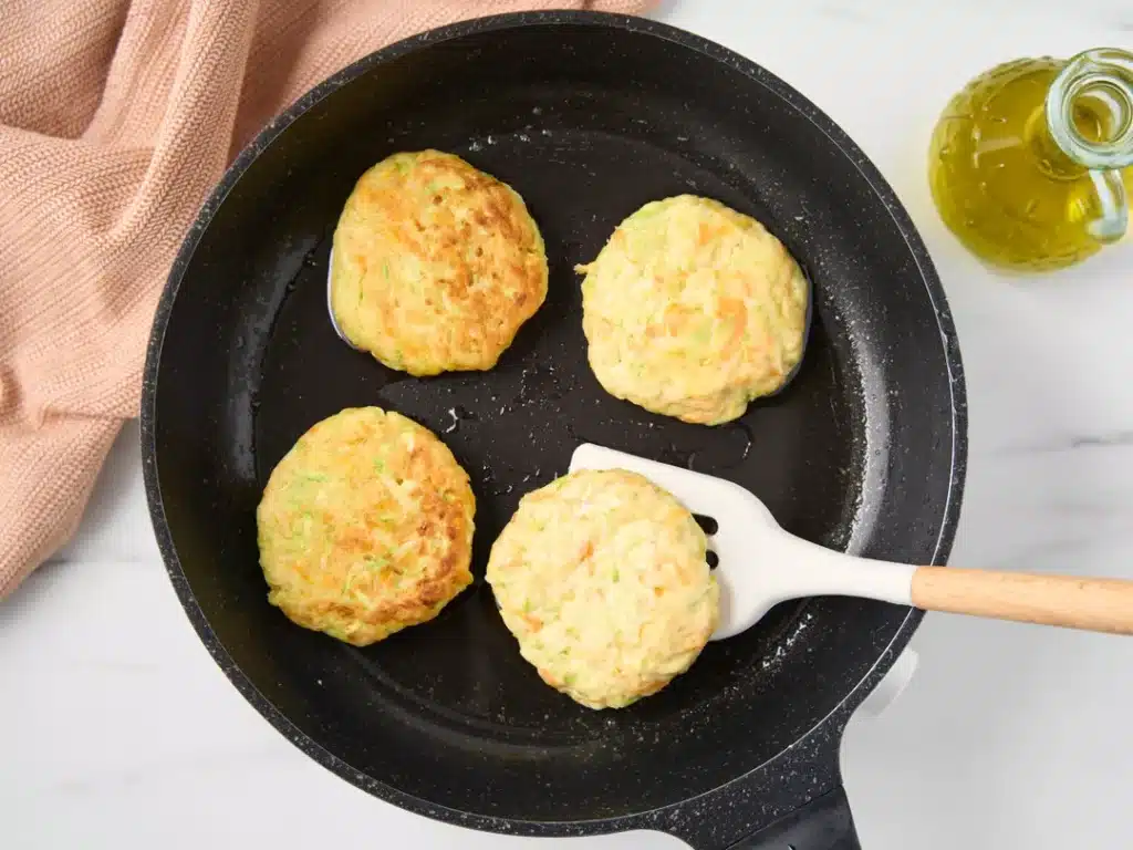 Four vegetable fritters frying in a black pan on a white surface with a spatula and olive oil bottle nearby.