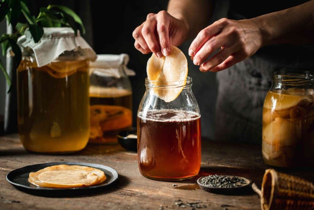 Hands making kombucha with SCOBY in a jar on a rustic wooden table with tea leaves nearby.