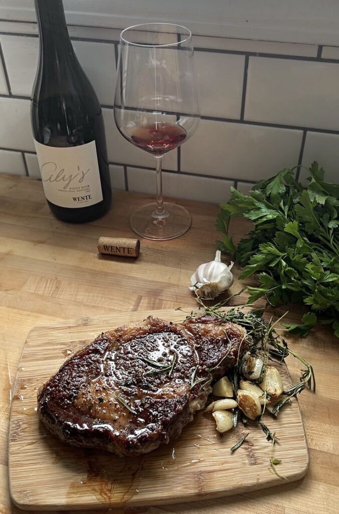 Seared steak with herbs and garlic on a board, paired with red wine, glass, and fresh parsley on wooden counter.