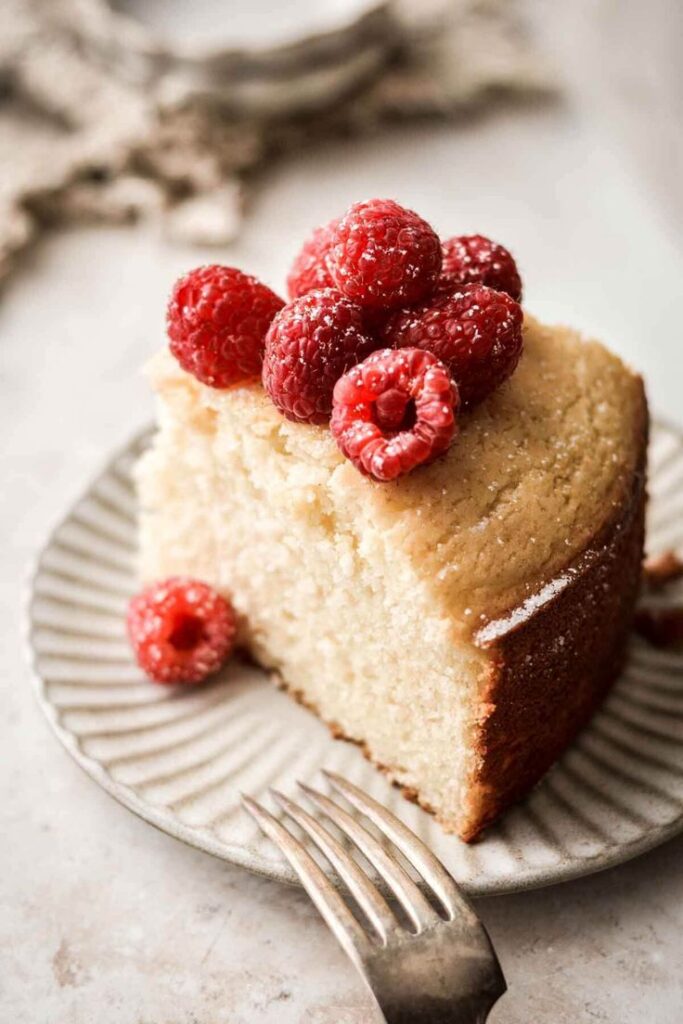 A slice of vanilla cake topped with fresh raspberries on a plate, with a fork beside it.