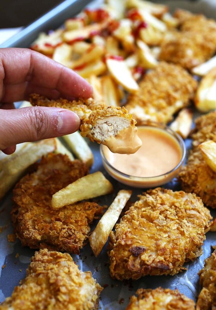 Hand dipping crispy chicken tender in sauce with fries on a tray. Perfect snack or appetizer.