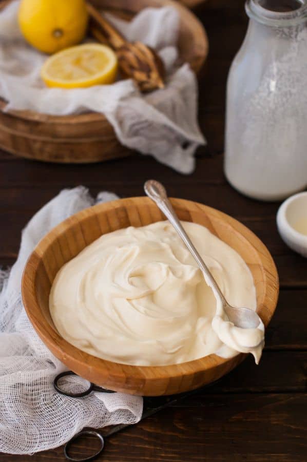 Creamy homemade mayonnaise in a wooden bowl with a spoon, surrounded by lemons and ingredients on a rustic table.