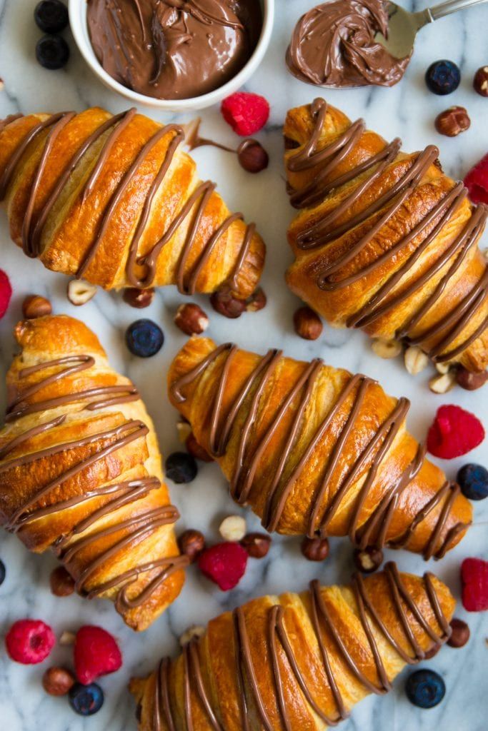 Chocolate-drizzled croissants with fresh berries and hazelnuts on a marble background, next to a bowl of chocolate spread.