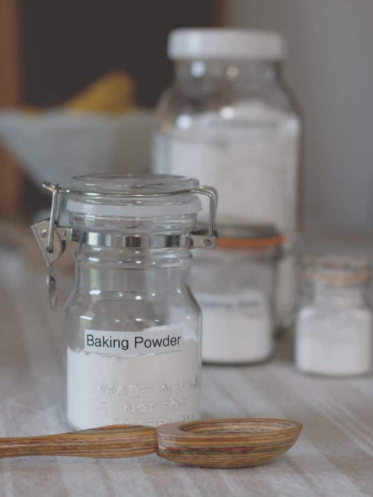 Glass jar of baking powder on a kitchen counter with a wooden spoon in front.