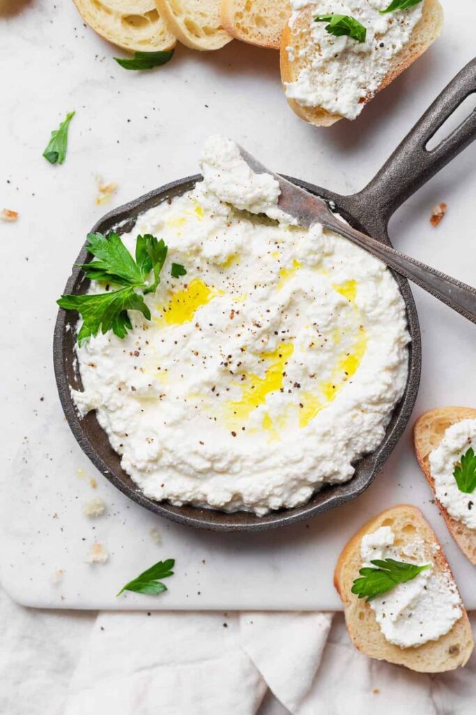 Creamy ricotta cheese spread on bread with parsley garnish, served in a pan, perfect for appetizers.