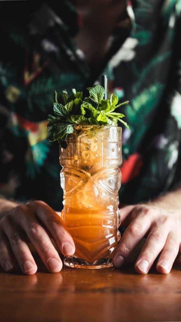 Tropical cocktail in a tiki glass garnished with fresh mint, held by a person wearing a floral shirt.