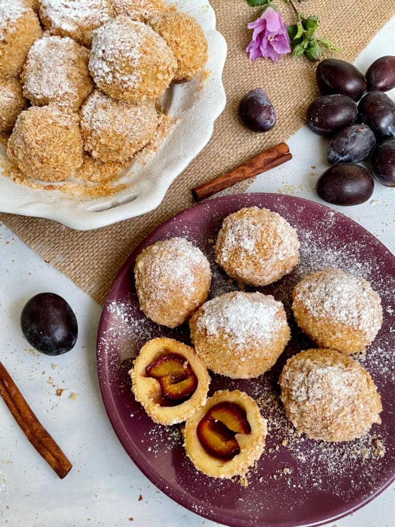 Plum dumplings sprinkled with powdered sugar on plates, surrounded by fresh plums and cinnamon sticks.