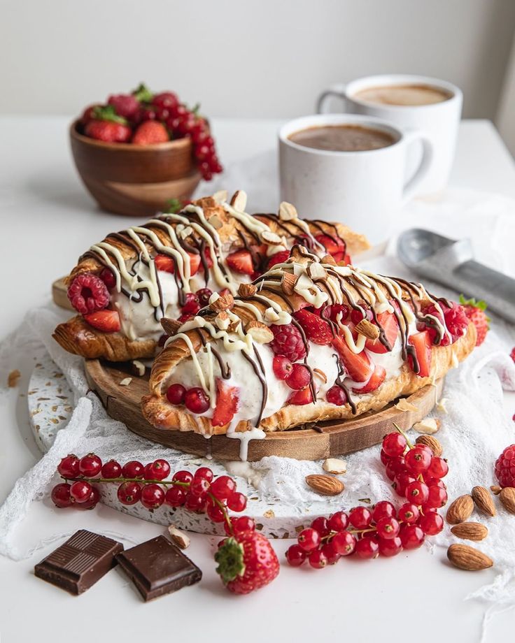 Delicious croissants topped with berries, cream, and chocolate drizzle, served with coffee and fresh fruit on a white table.