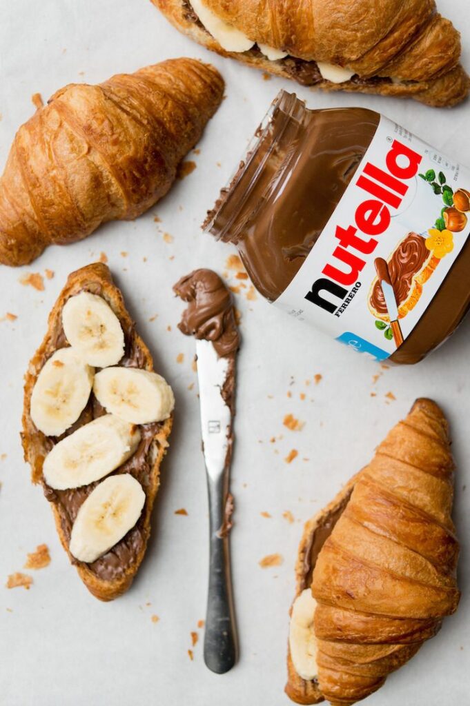 Croissants with Nutella and banana slices, surrounded by a jar of Nutella and a knife on a white surface.