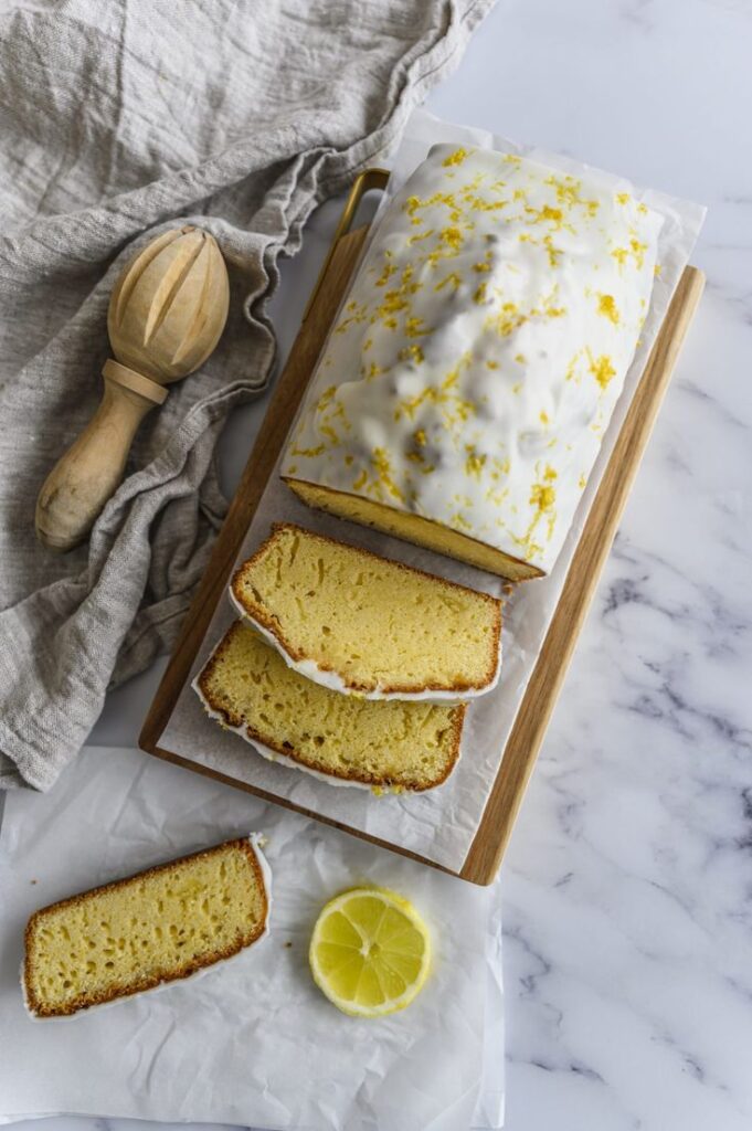 Lemon drizzle loaf with icing, sliced on a wooden board. Linen cloth and lemon wedge on marble countertop.