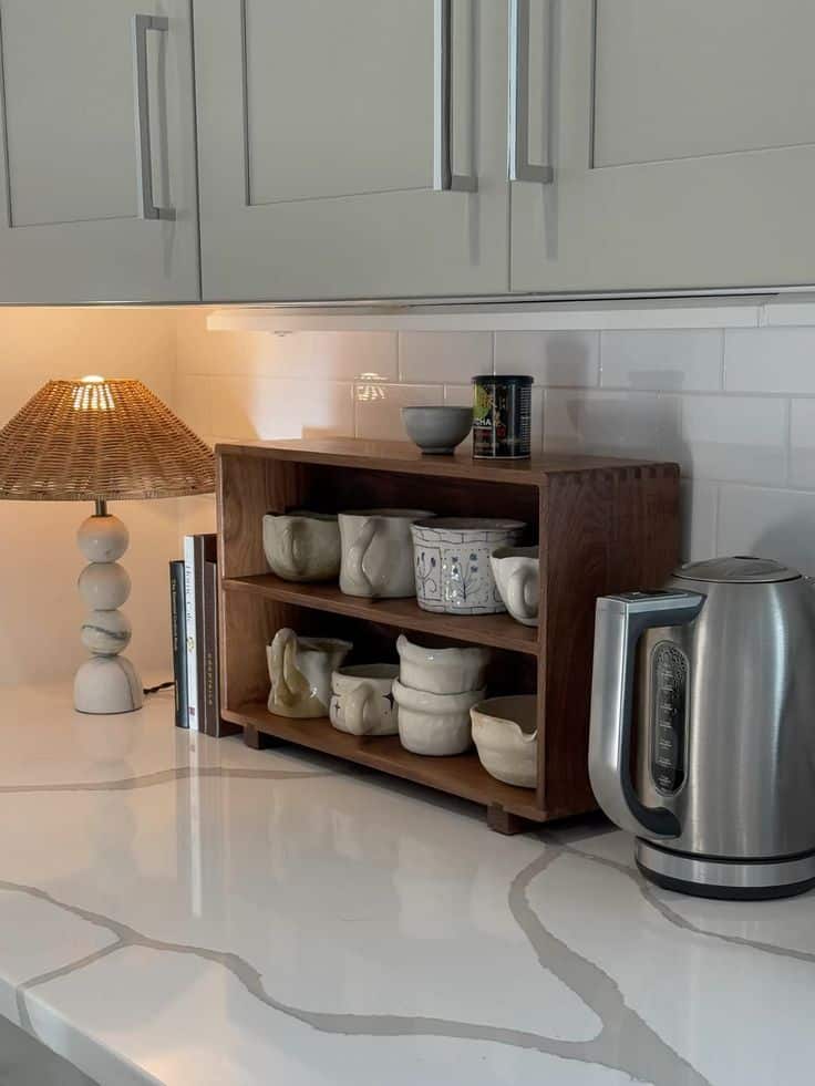 Modern kitchen counter with a wooden shelf, ceramic cups, books, an electric kettle, and a stylish lamp.