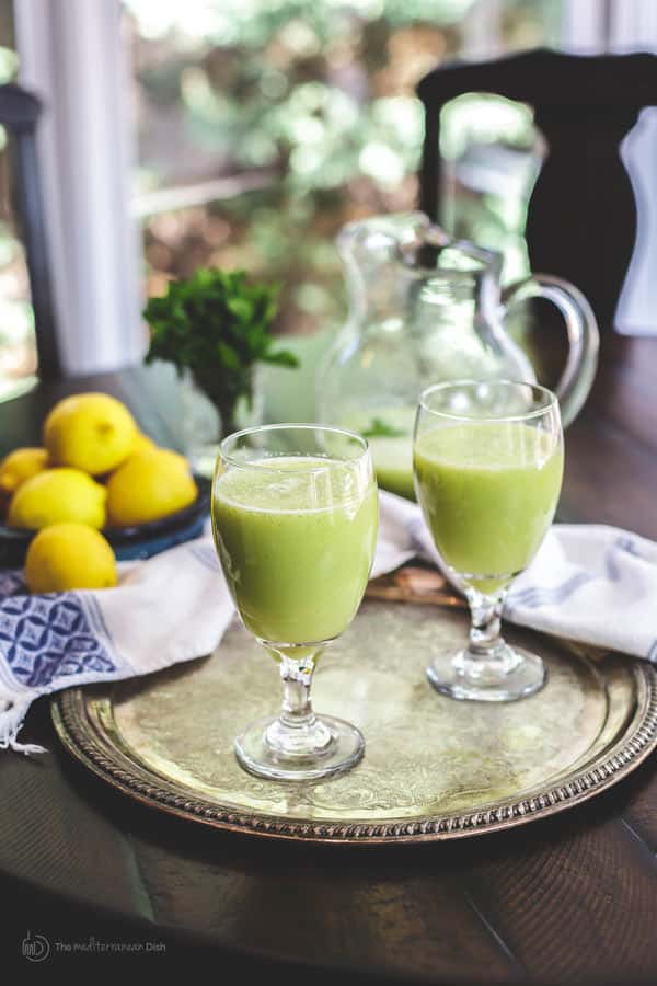 Two glasses of green smoothie on a tray, with lemons and parsley in the background. Refreshing drink setting.