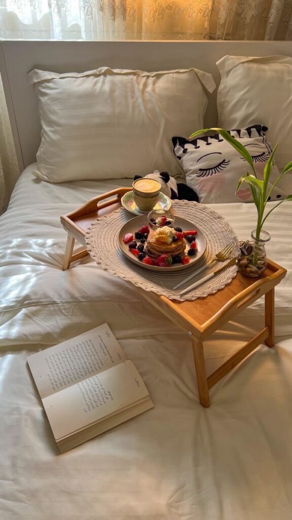 Breakfast in bed with pancakes and berries on a tray, accompanied by coffee and an open book in a cozy setting.