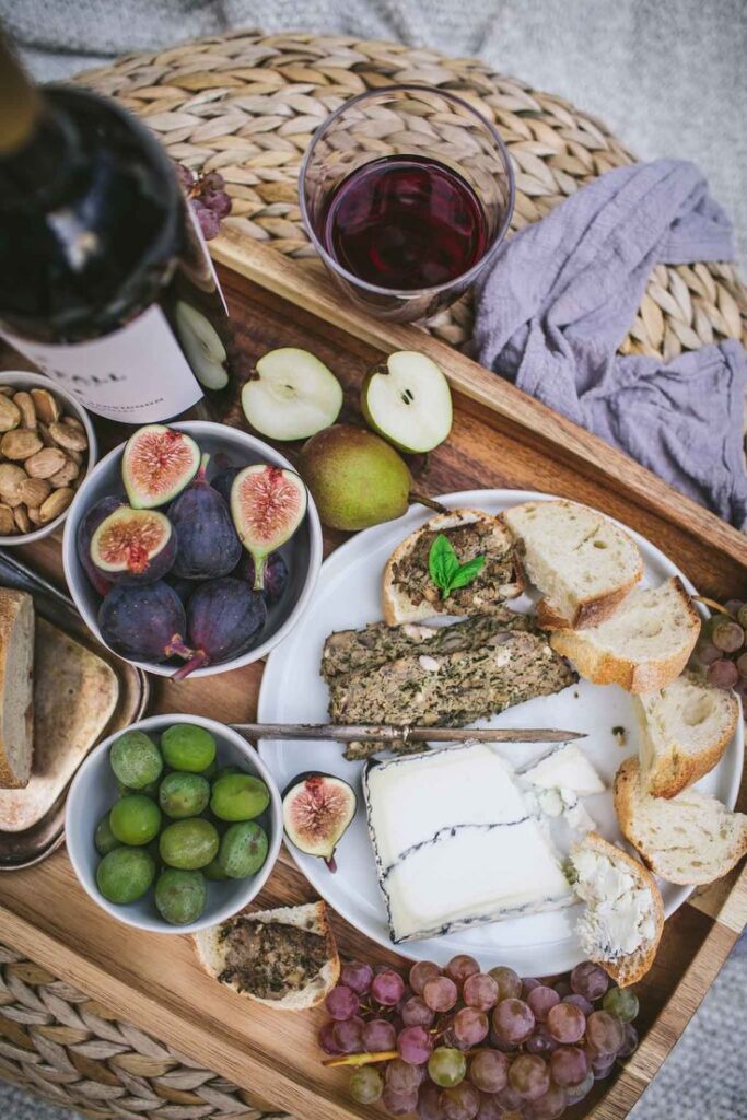 Cheese board with figs, grapes, nuts, bread, and red wine on a wooden tray. Perfect for a gourmet picnic setting.