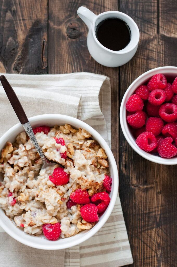 Bowl of oatmeal topped with fresh raspberries and walnuts, with a side of raspberries and a pour of syrup on a rustic table.