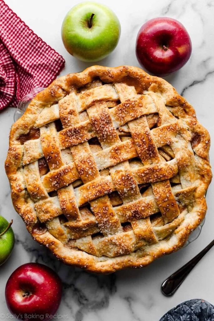 Homemade lattice apple pie on marble with fresh apples and red checkered cloth.