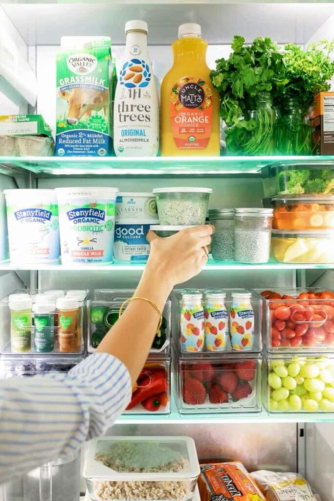 Open fridge with organized organic groceries, drinks, and fresh produce, as a hand reaches for a container.