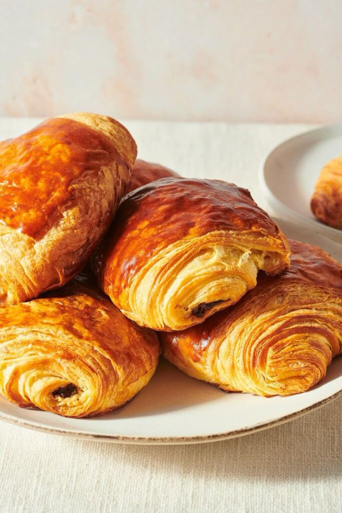 Plate of flaky, golden-brown pain au chocolat pastries on a white tablecloth, perfect for a delicious breakfast treat.